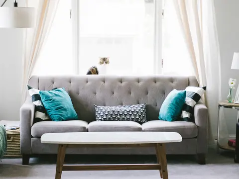 A grey sofa dressed with teal and patterned cushions and a folded throw beside a bright window.