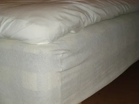 A quilted white mattress protector fitted over the corner of a mattress on a bed base.