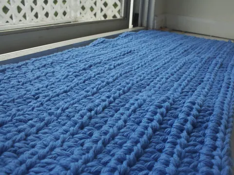 A blue crocheted cotton rug lying on a tiled floor beside a window.