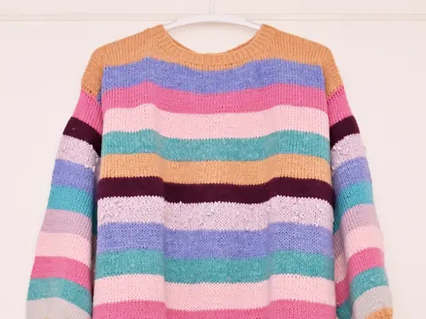 A hand-knitted pullover with wide stripes in peach, lilac, pink, teal and grey hanging on a white wall.