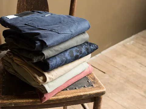 A stack of folded chino trousers in navy, grey, camouflage, tan, sage and rust resting on an old wooden chair.
