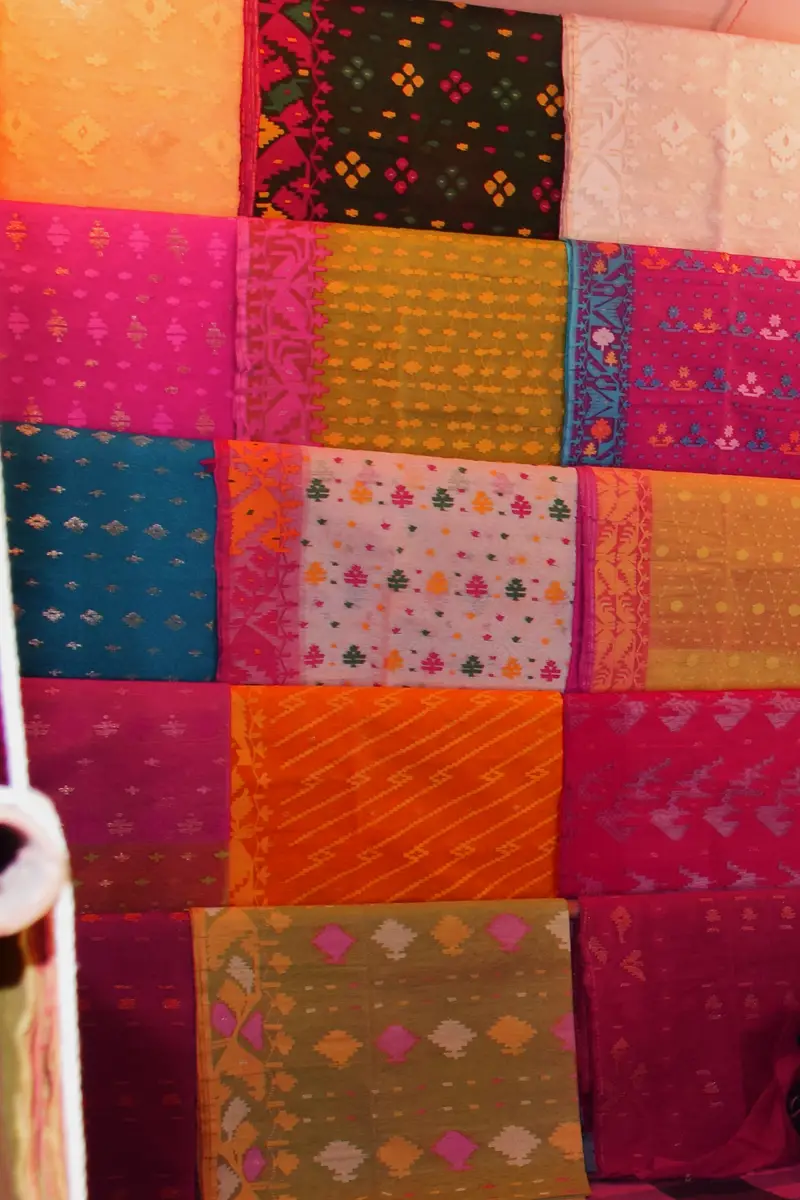 A wall of folded jamdani sarees in pink, orange, teal and gold hanging in rows inside a shop in Sonargaon, Bangladesh.