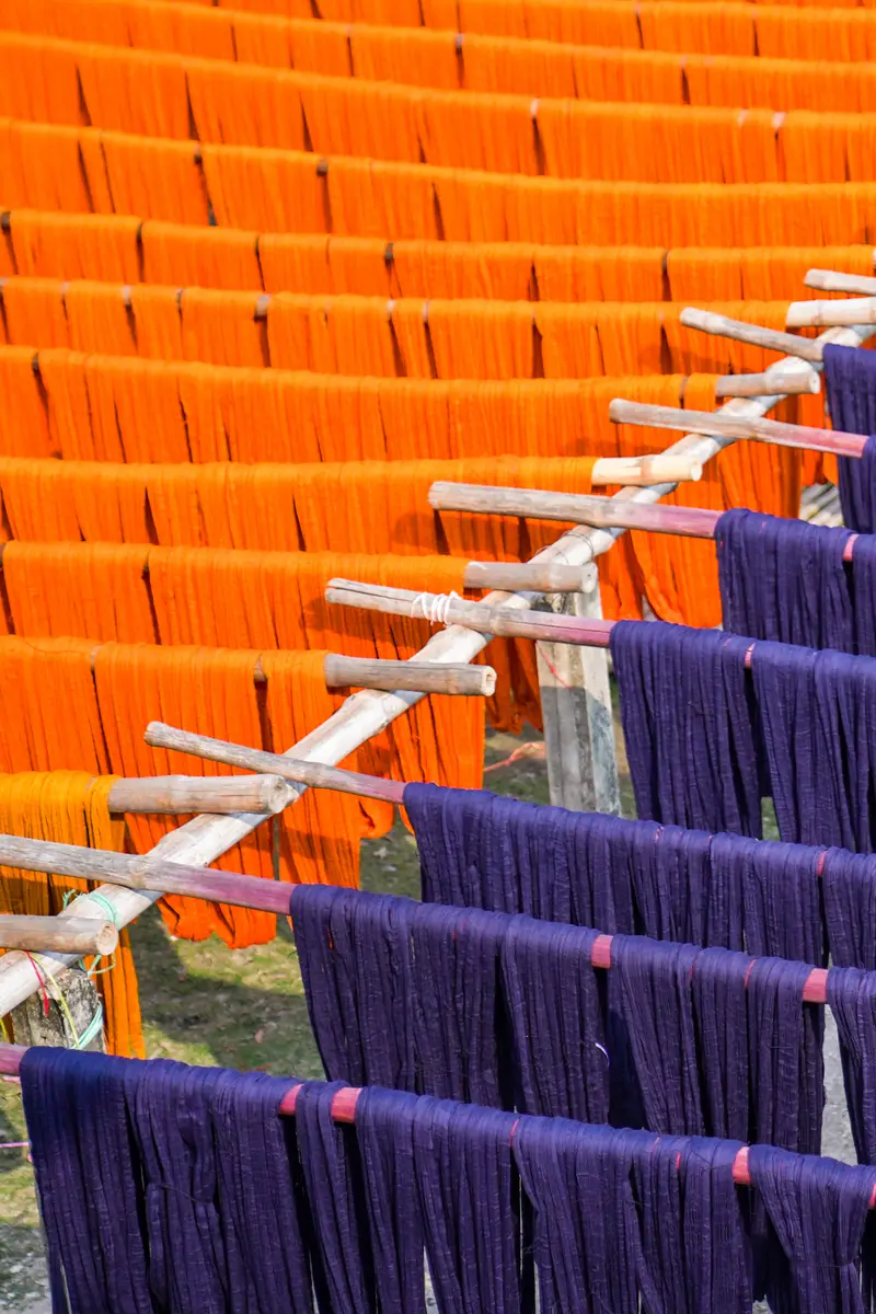 Hanks of freshly dyed yarn in deep indigo-purple and bright orange hanging from bamboo poles to dry in the sun.