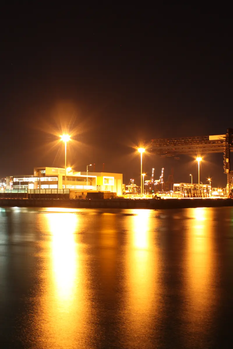 A container terminal at night with its tall yellow floodlights and quay cranes reflected as long golden streaks across dark water.