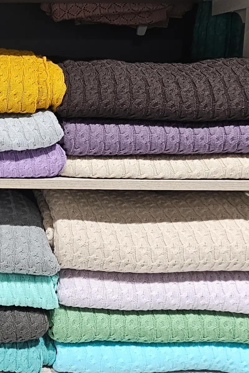 Folded knitted sweaters stacked by colour on light wooden shelves.