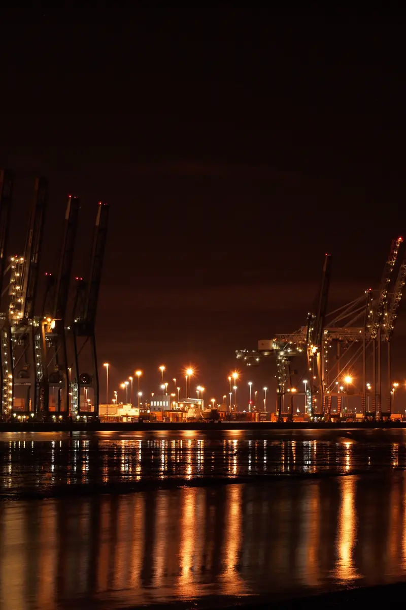 Container cranes lit up along a dock at night, reflected in still water.