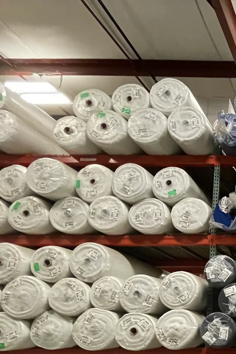 Rolls of white, blue and grey fabric stacked on red warehouse racking, each roll wrapped in plastic with a small ticket.