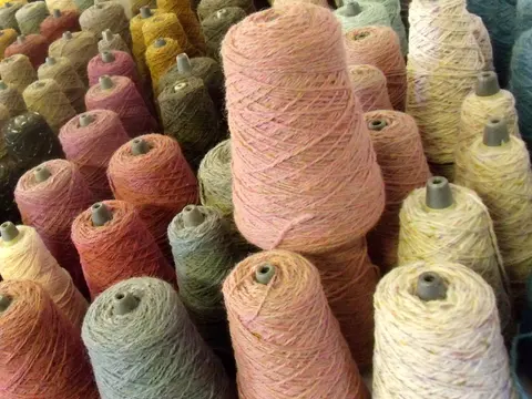 Rows of yarn cones in muted melange shades of pink, grey, green and cream on a mill floor.