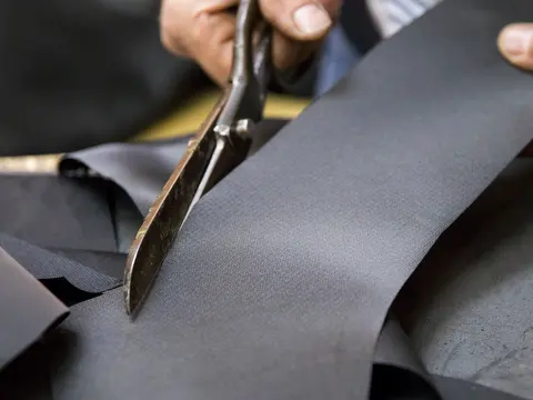 Hands guiding tailoring shears through a length of grey suiting cloth.