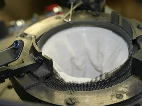 Close view of a circular knitting machine needle cylinder with the white knitted fabric tube forming inside.