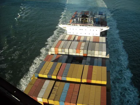Stacked shipping containers on the deck of a cargo ship seen from above as it leaves a wake at sea.